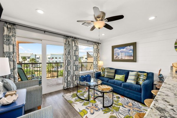 Coastal living room with blue sofa, floral rug, and glass coffee tables facing sliding doors to a balcony.