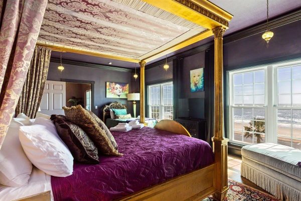 Luxurious gold four-poster bed with a purple quilt and plush pillows in a dark, seaside bedroom.